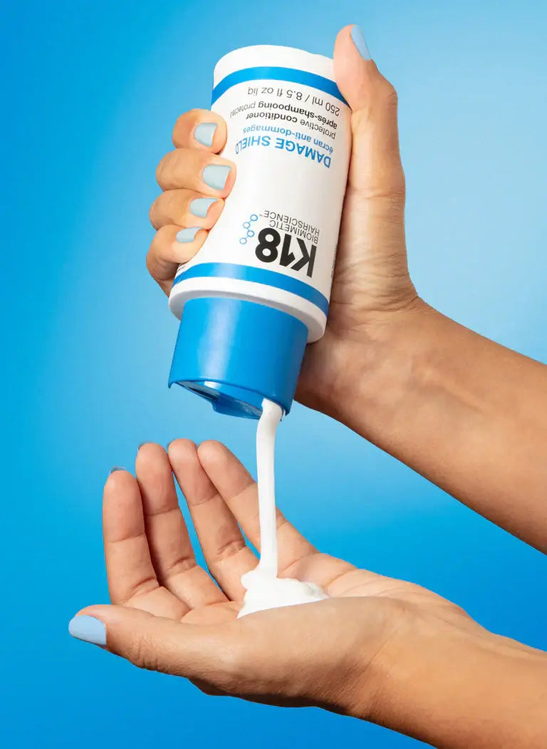 Hands using DAMAGE SHIELD Protective Conditioner with K18PEPTIDE against blue background, enhancing hair protection and shine.