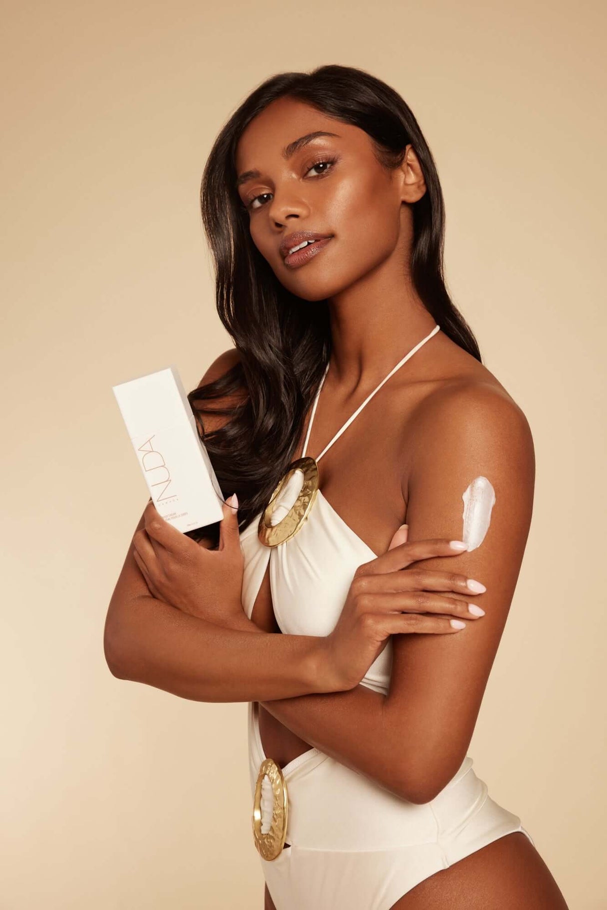 Woman applying body cream in a beige swimsuit while holding skin care product box.