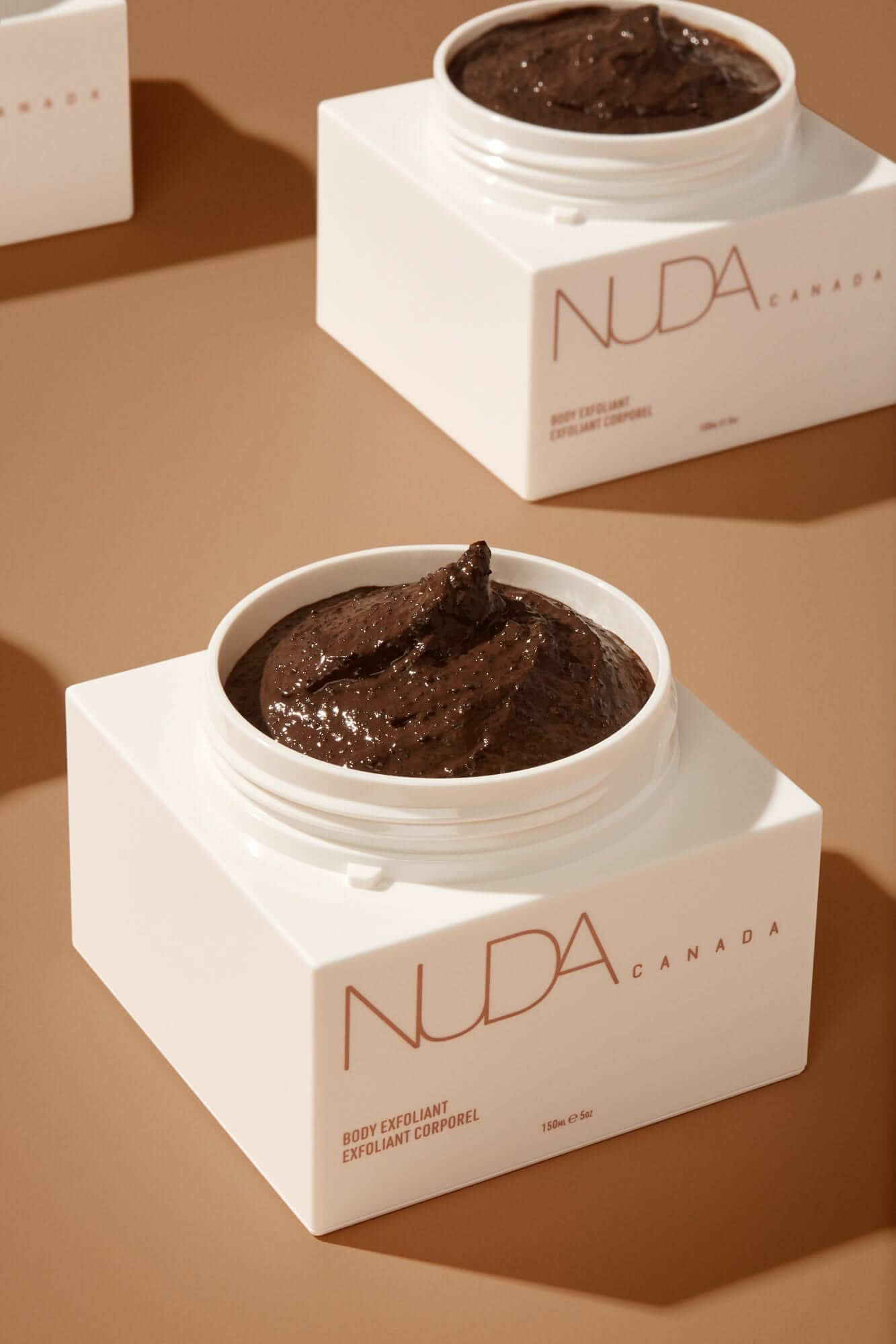 Close-up of NUDA Canada body exfoliant jar on a brown background, showcasing rich texture for skincare and tanning preparation.