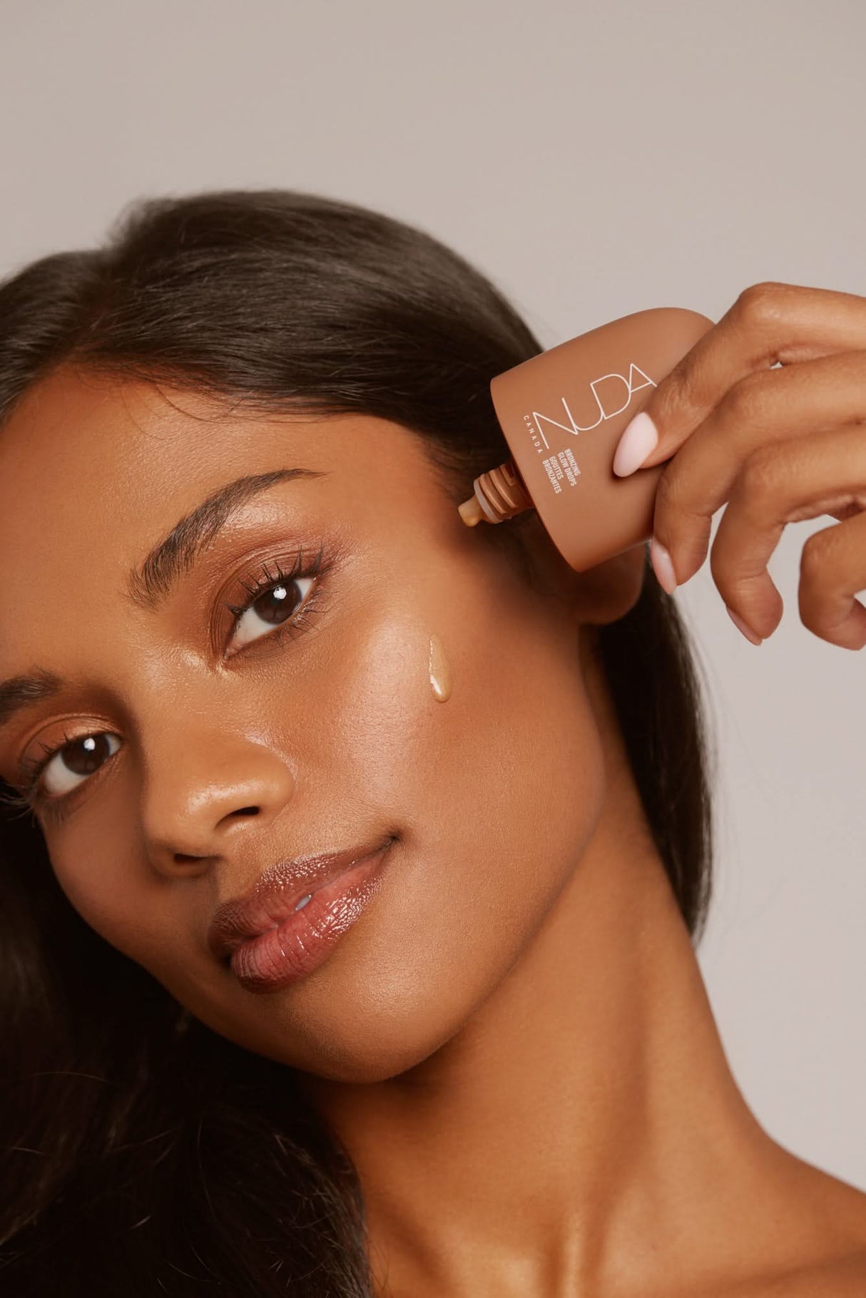 Woman applies Nuda Let It Glow Bronzing Glow Drops to achieve a sun-kissed glow. Ideal for beauty enthusiasts and holiday gifting.