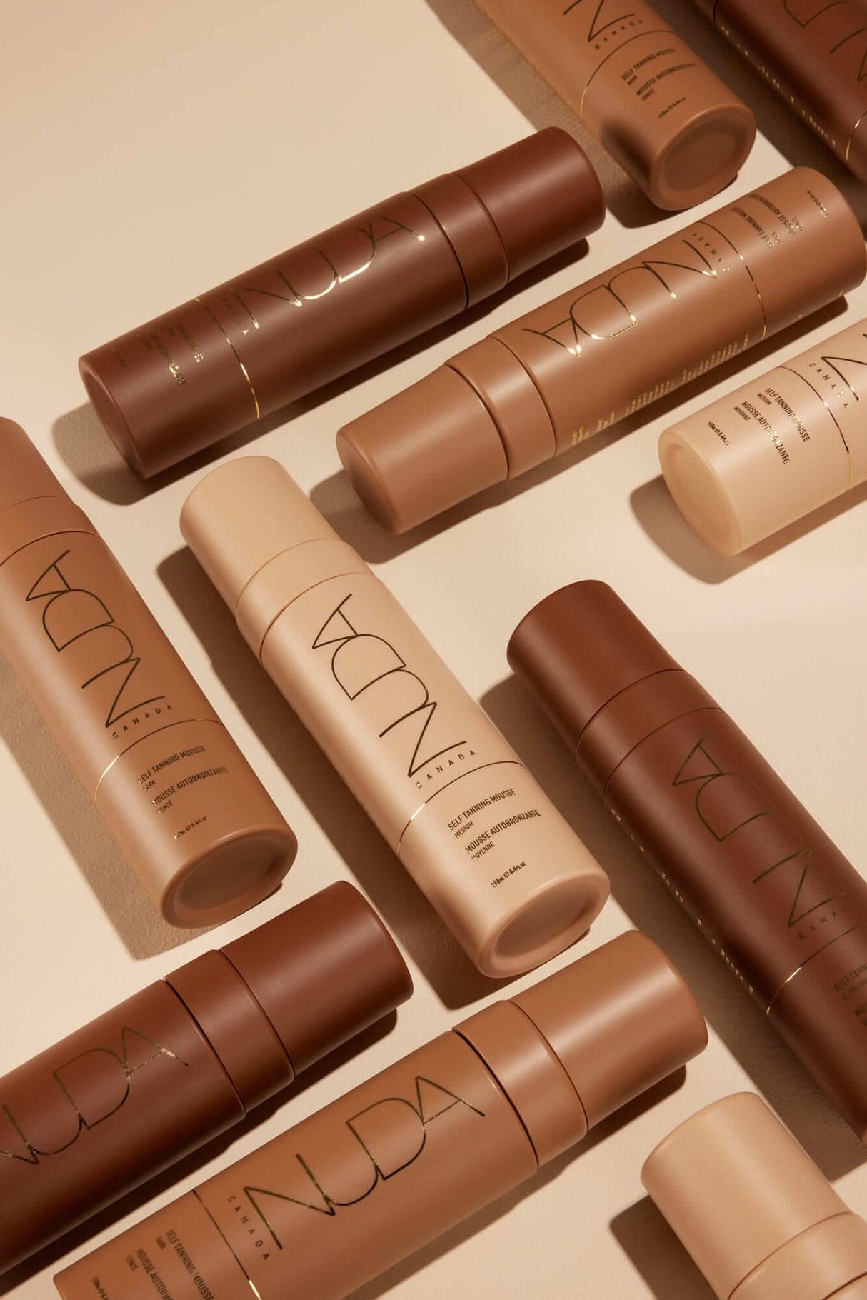 Various shades of self-tanning mousse bottles and tanning products for a radiant glow on a neutral background.