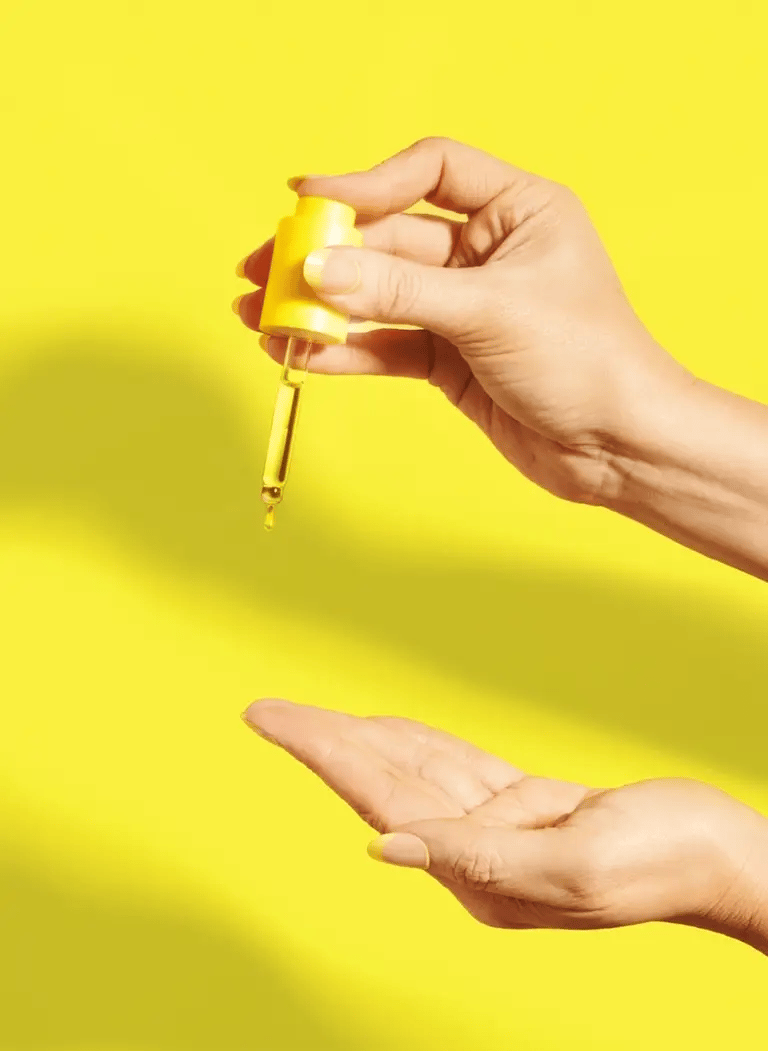 Hand applying Molecular Repair Hair Oil dropper against yellow background.