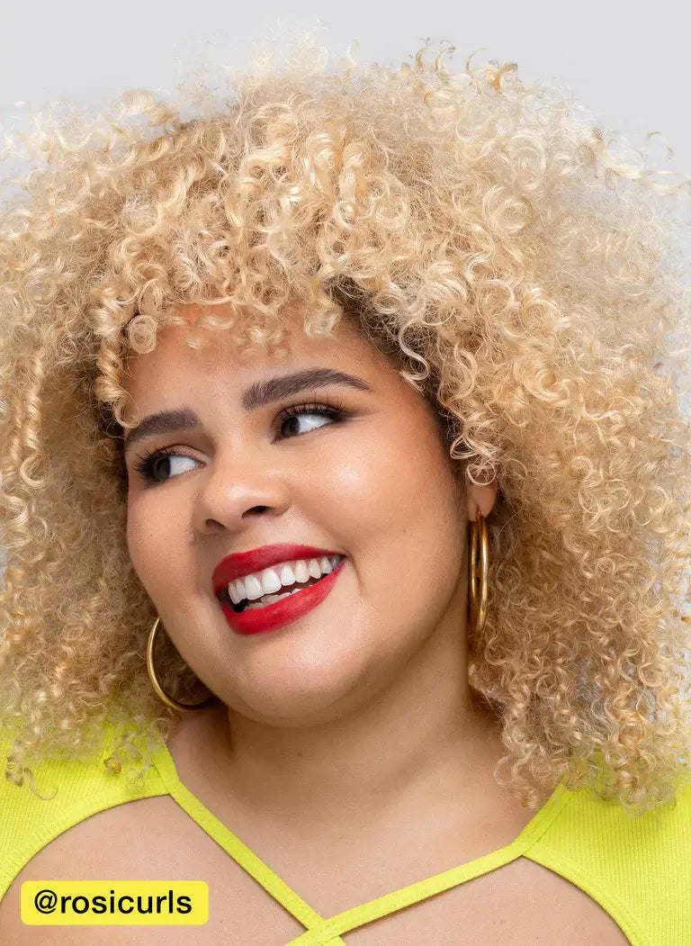 Woman with curly, blonde hair smiles wearing yellow top and hoop earrings.