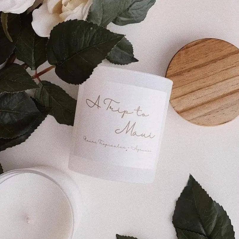 Candle labeled 'A Trip To Maui' surrounded by green leaves, featuring tropical floral and citrus fragrance in a glass jar with a wooden lid.