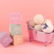 Stacked boxes and bath bombs in a mini tub against pink background, showcasing all-natural ingredients for soothing skincare routine.