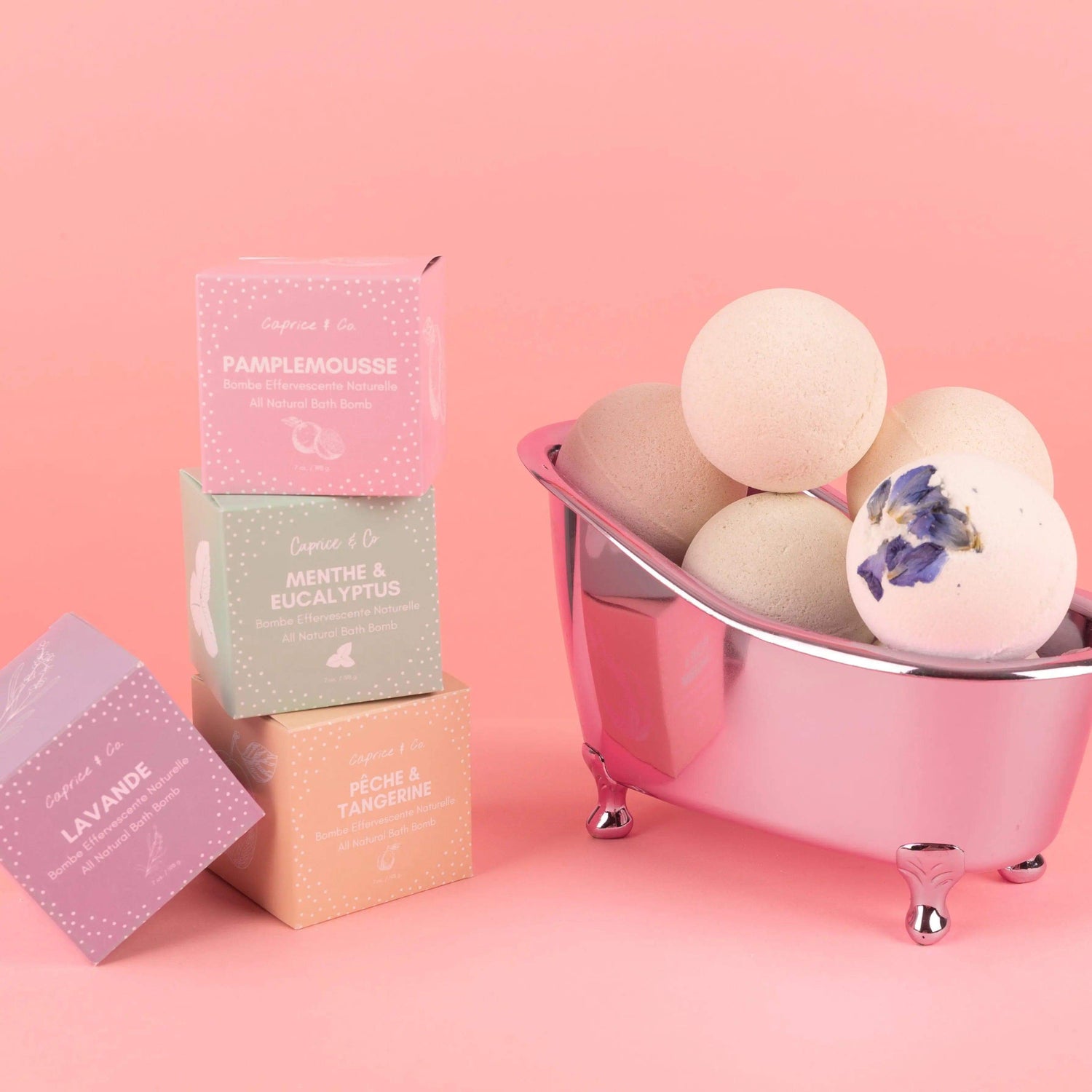 Stacked boxes and bath bombs in a mini tub against pink background, showcasing all-natural ingredients for soothing skincare routine.
