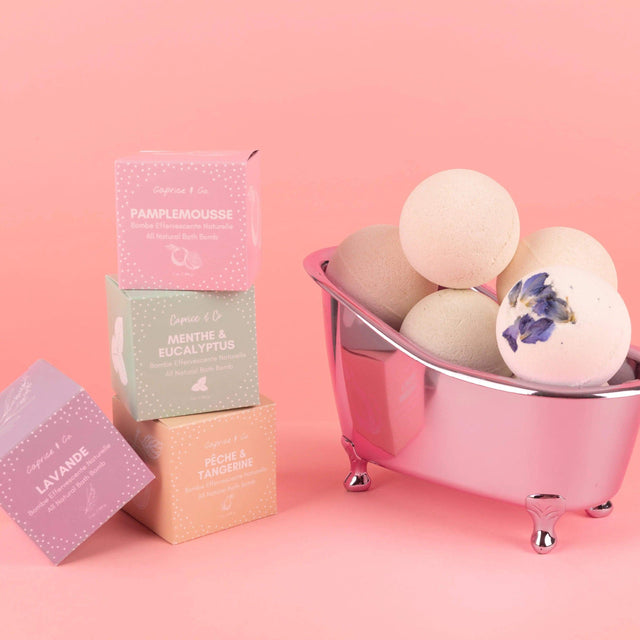 Stacked boxes and bath bombs in a mini tub against pink background, showcasing all-natural ingredients for soothing skincare routine.