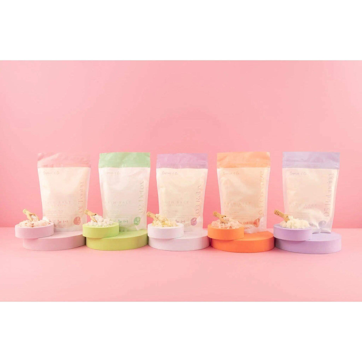 Colorful packages of all-natural bath salts with essential oils displayed against a pink background.