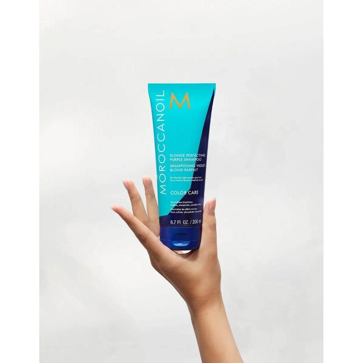 Hand holding Moroccanoil Blonde Perfecting Purple Shampoo for brassiness elimination.
