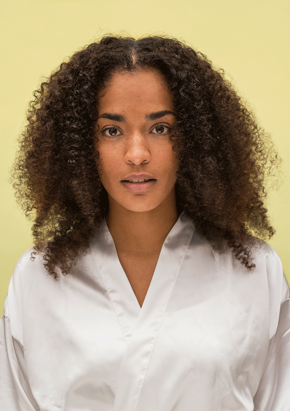 Woman with defined curly hair using BOUNCE.ME Curl Glitz Kit for hydrated and frizz-free curls against a yellow background.