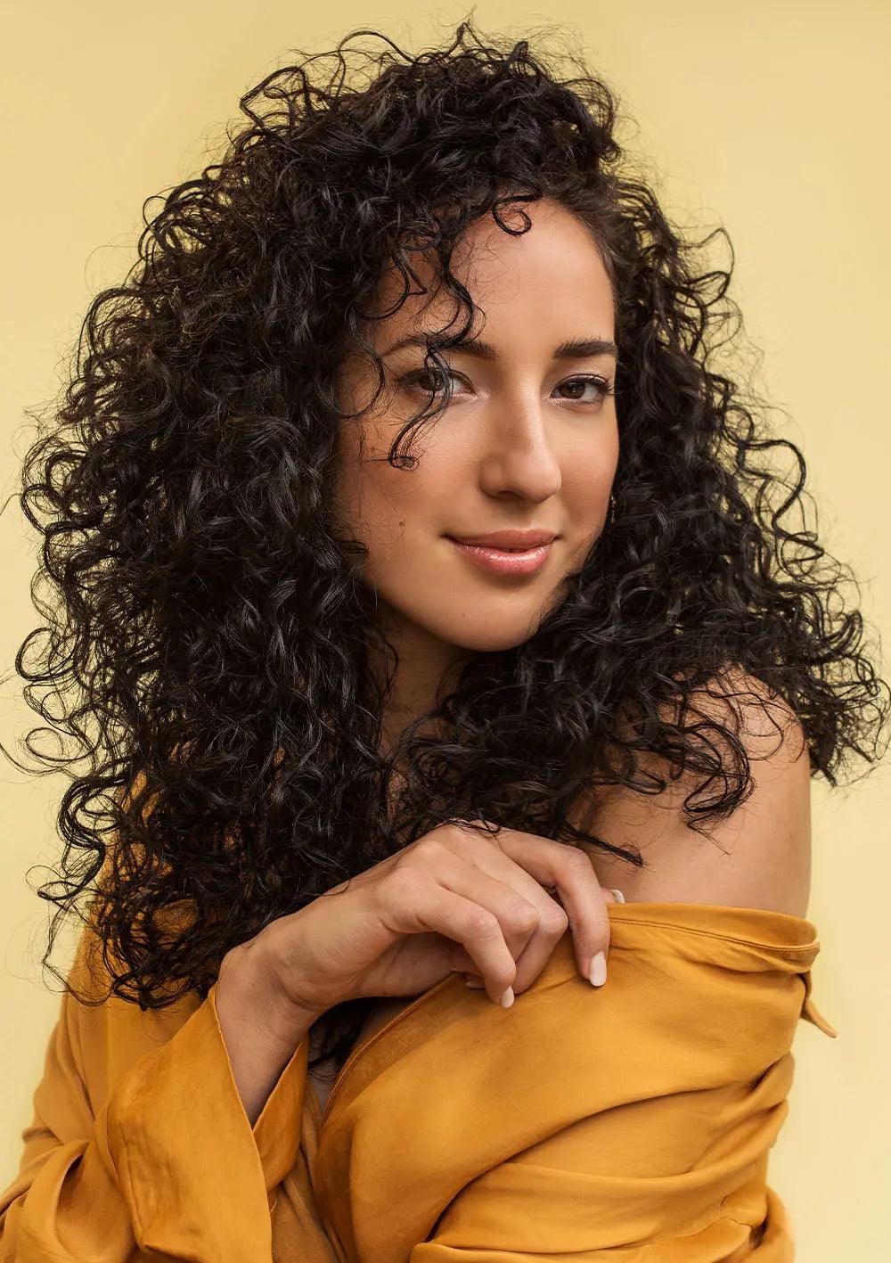 Person with voluminous curly hair wearing a mustard-yellow top on a yellow background.