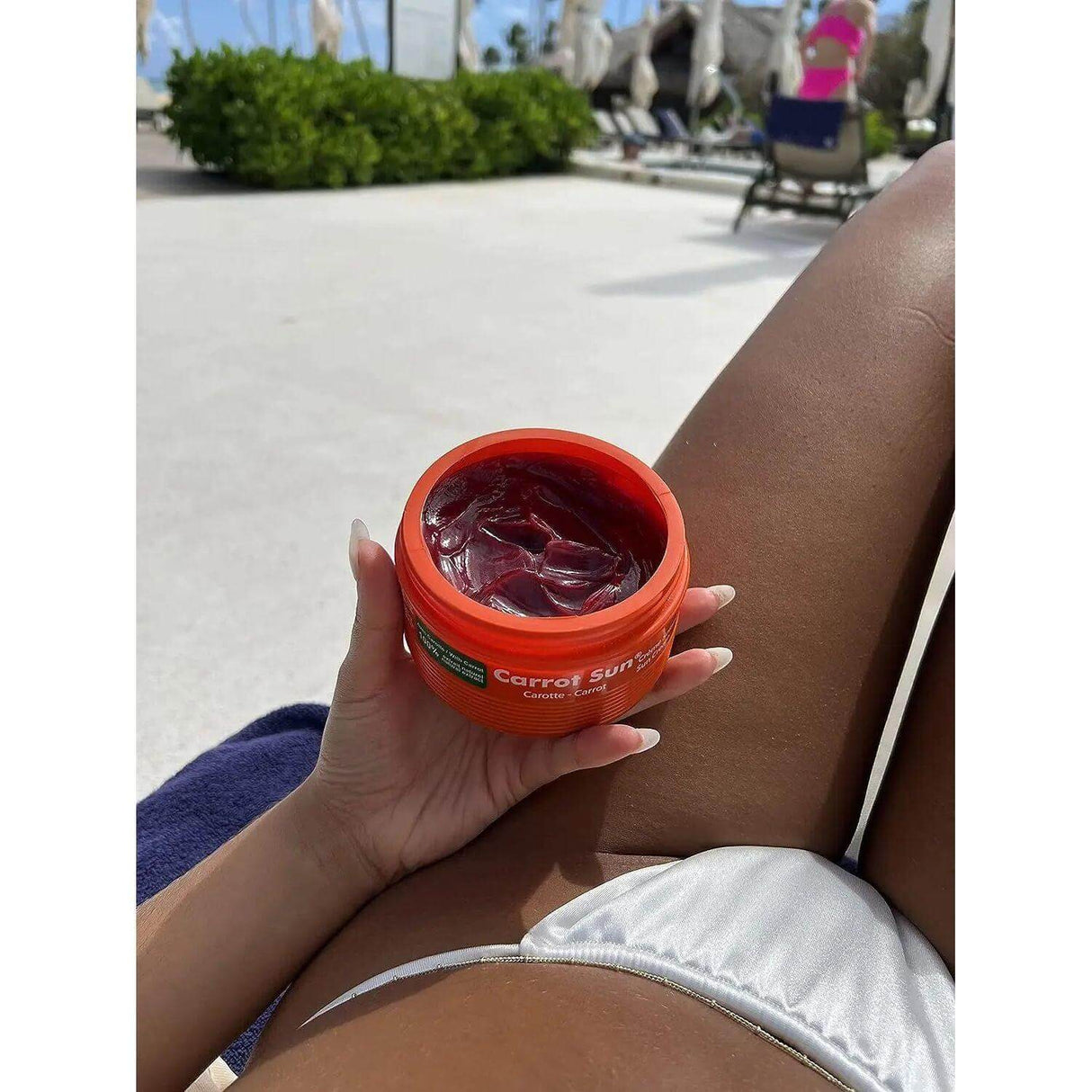 Person holding Carrot Cream tanning formula by a pool, promoting dark long-lasting tan with carrot oil and L-Tyrosine.