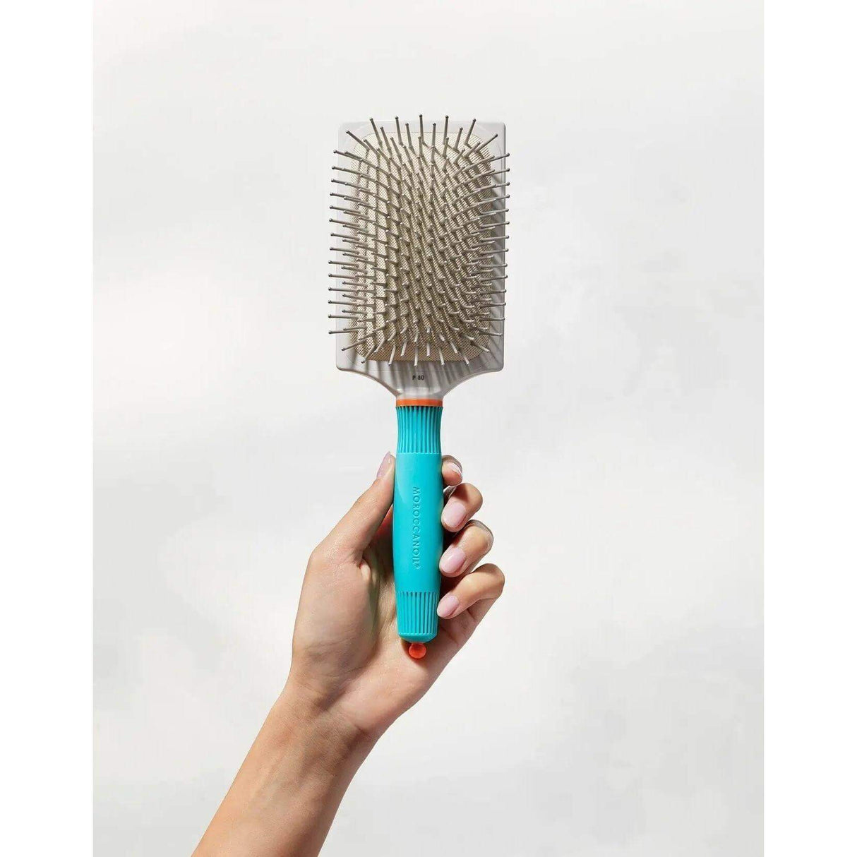 Hand holding Moroccanoil Ceramic Paddle Brush with turquoise handle, promoting shine and reducing drying time.