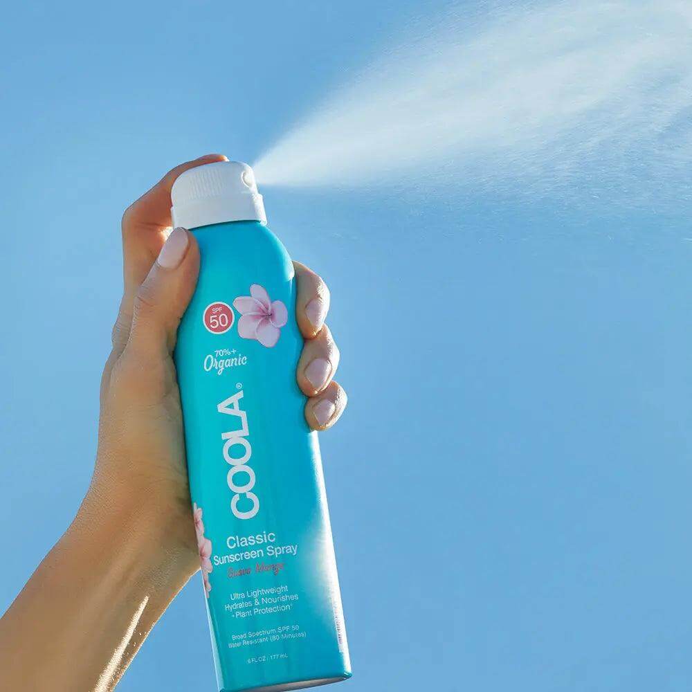 Hand holding Classic Body Organic Sunscreen Spray SPF 50 in front of blue sky, nozzle spraying fine mist.