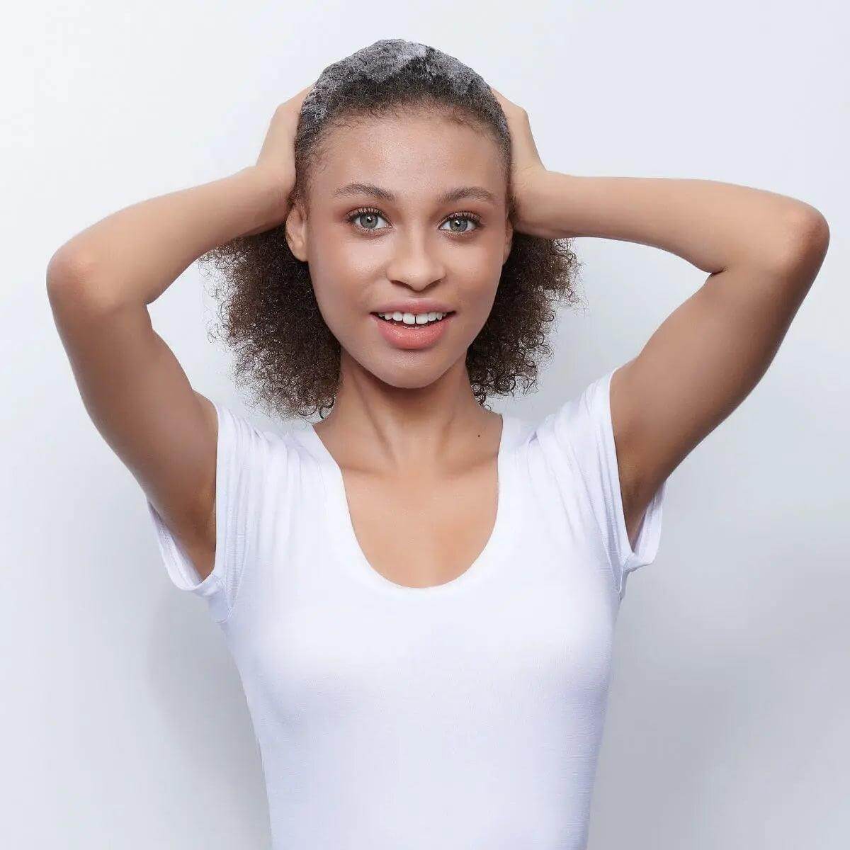 Woman smiling with curly hair styled using Moroccanoil Curl Control Mousse, wearing a white shirt, holding hair with both hands.