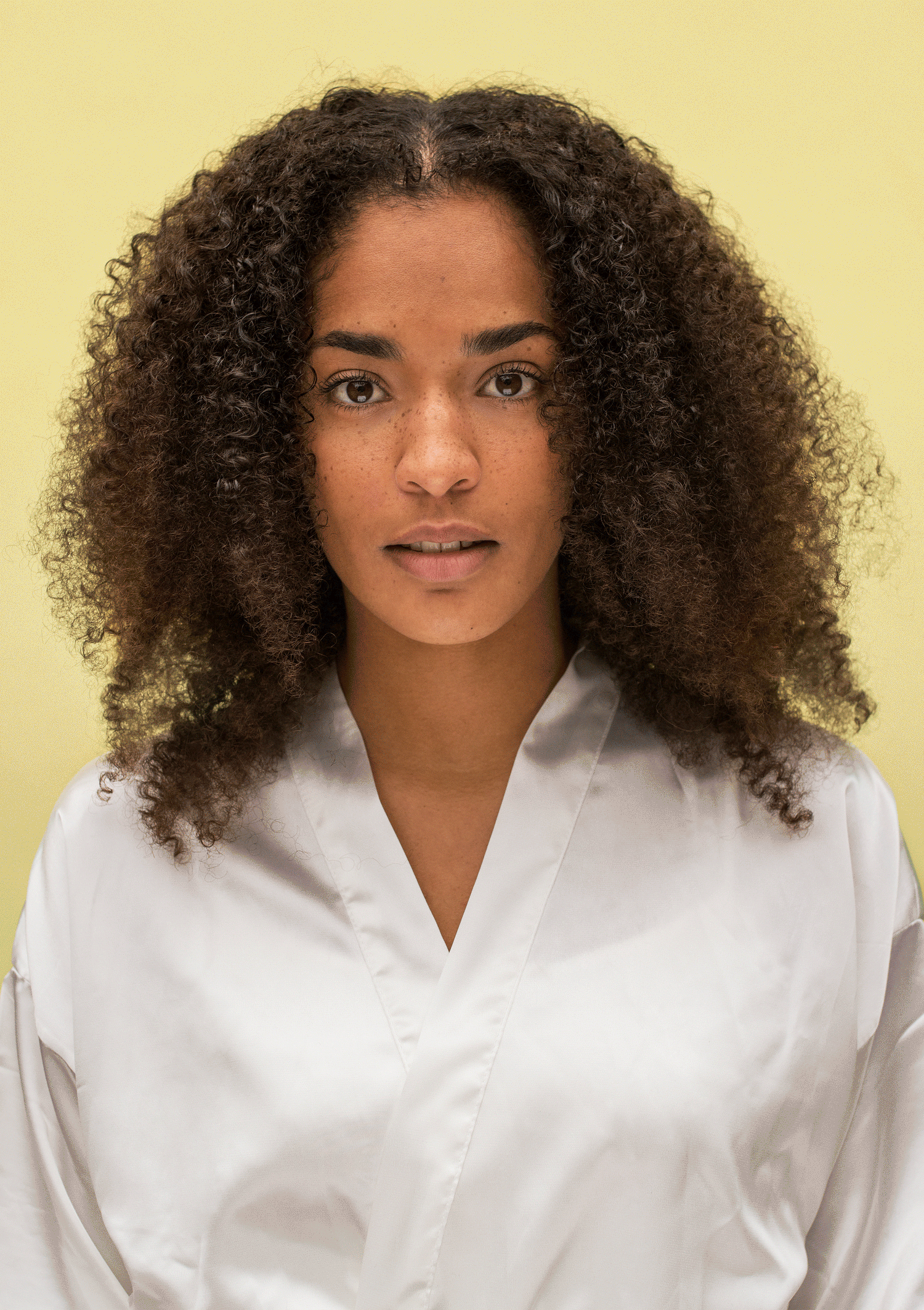 Person with curly hair wearing a white robe, standing against a yellow background.