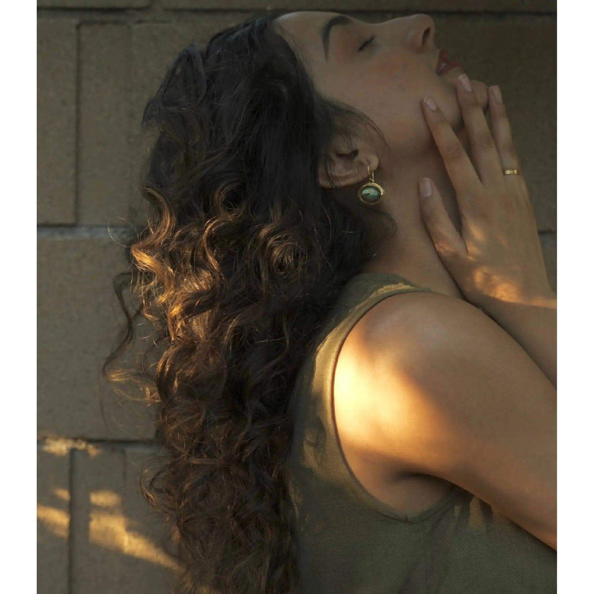 Woman with long, curly hair leaning back against a brick wall, showcasing defined curls with a golden glow.