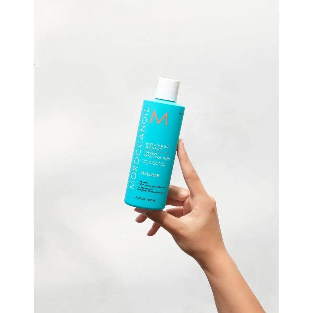 Hand holding Moroccanoil Extra Volume Shampoo bottle against white background.