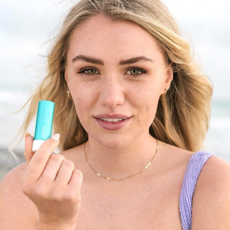 Woman holding Classic Liplux® Organic Lip Balm Sunscreen SPF 30 at the beach, showcasing its use for lip protection and hydration.