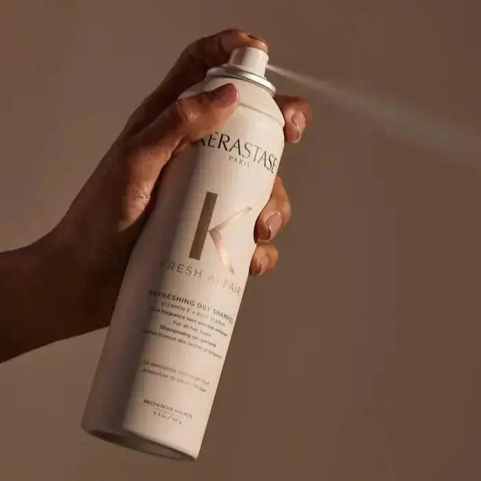 Hand holding a can of Kérastase Fresh Affair Oil-Absorbing Dry Shampoo spraying on brown background.