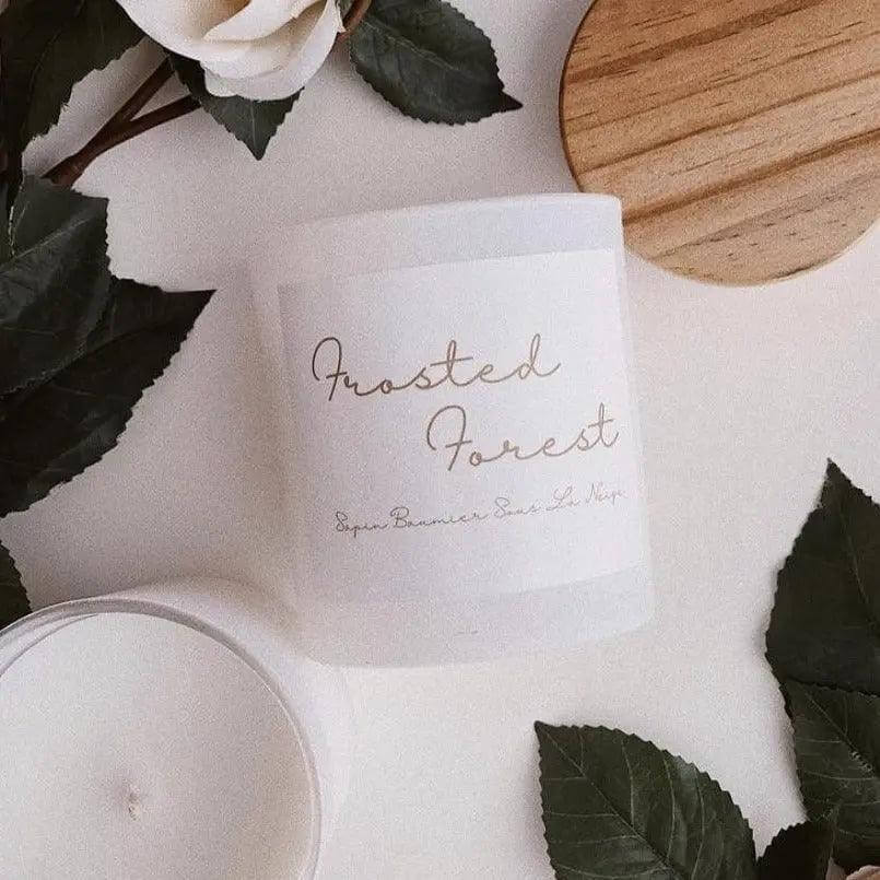 Frosted Forest candle with blue spruce and balsam fir scent, in a glass jar with a wooden lid, surrounded by green leaves.