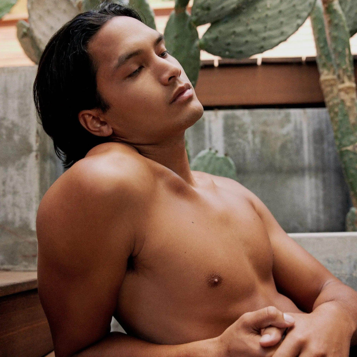 Person relaxing outdoors with eyes closed, leaning against a wall, surrounded by cacti.