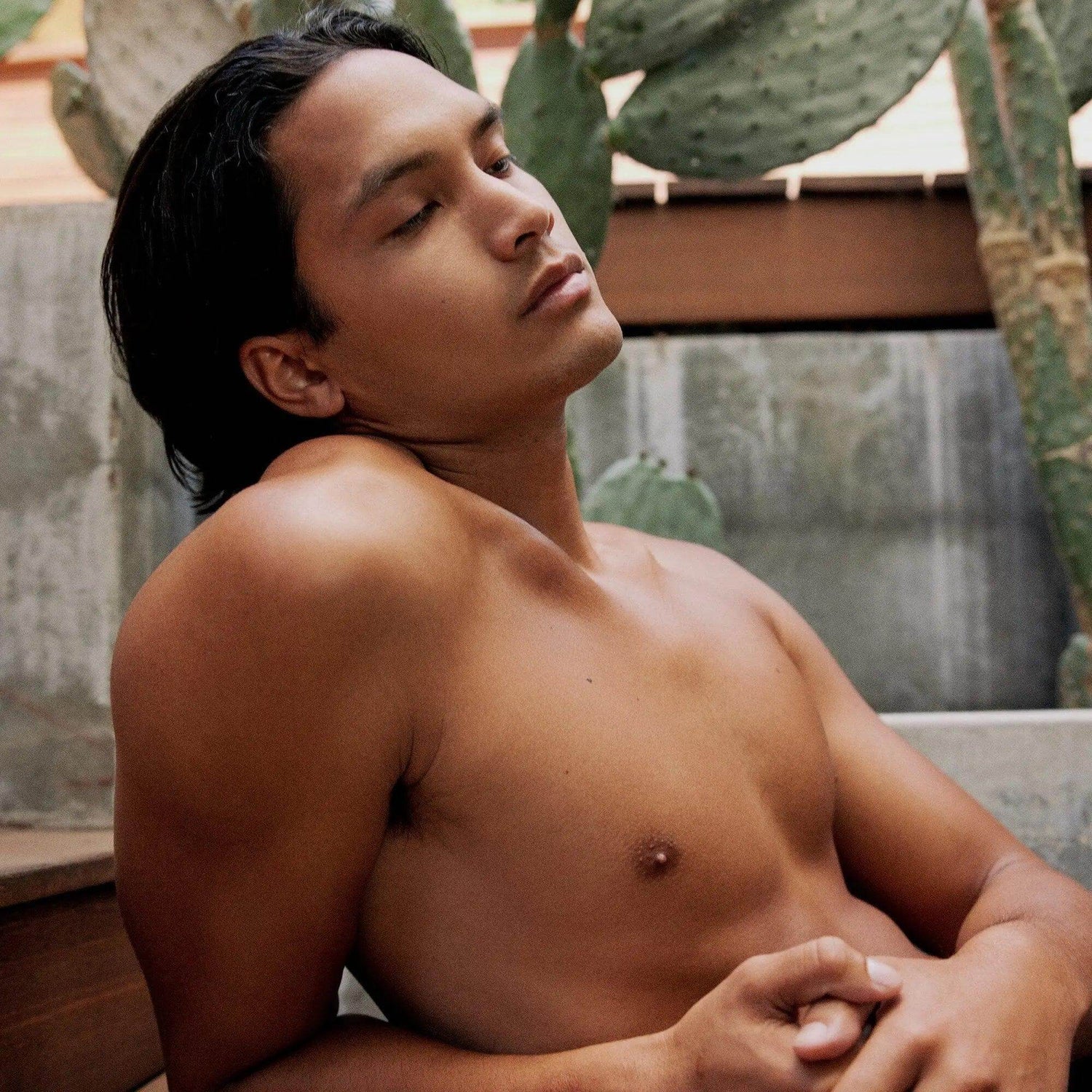 Person relaxing outdoors with eyes closed, leaning against a wall, surrounded by cacti.