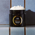 Gilded Woods Body Scrub jar on a tiled surface, featuring exfoliating and hydrating properties with pure cane sugar and coconut oil.