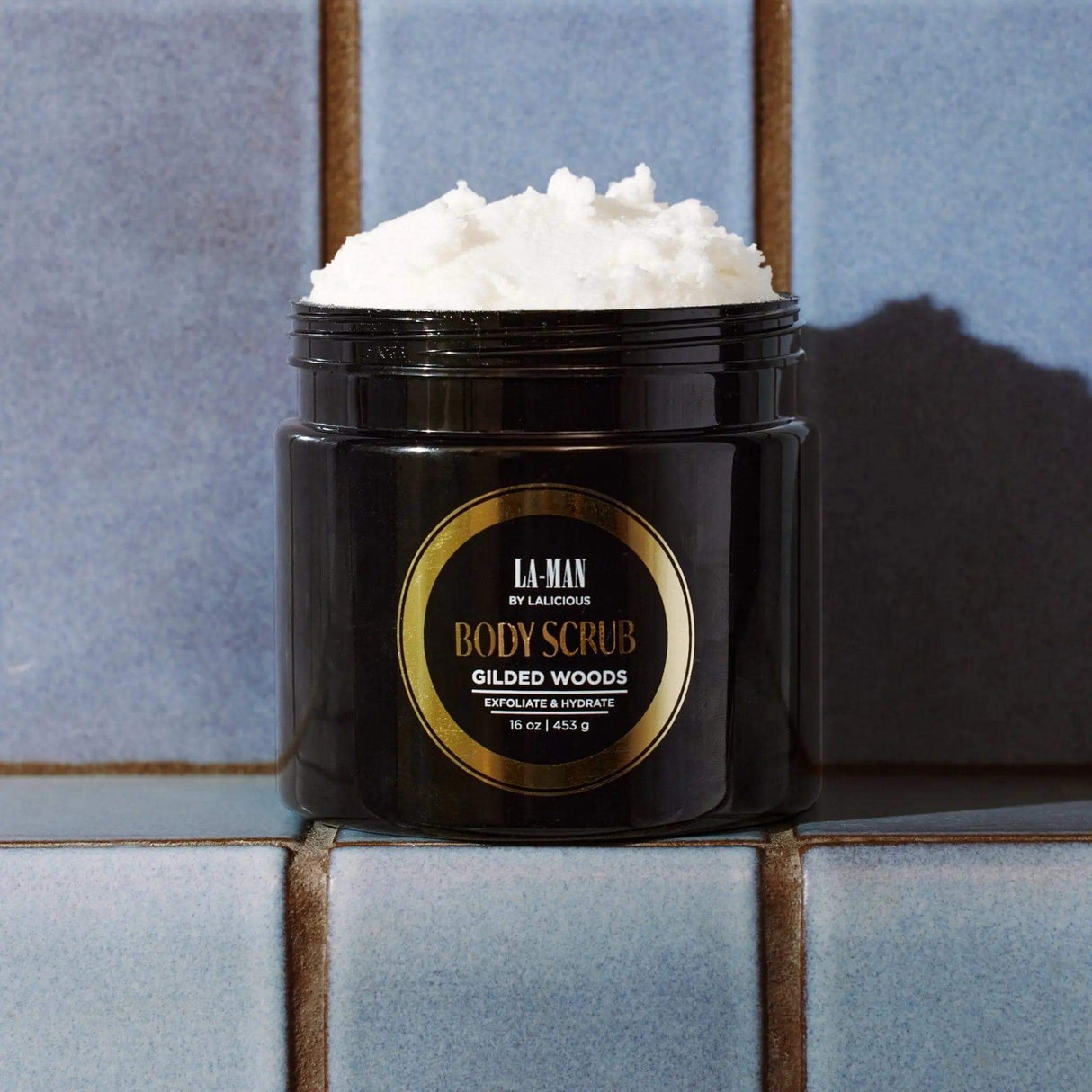 Gilded Woods Body Scrub jar on a tiled surface, featuring exfoliating and hydrating properties with pure cane sugar and coconut oil.