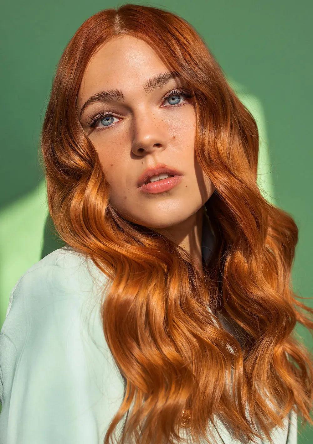 Person with long, wavy red hair against a green background, showcasing glossy and healthy hair texture.
