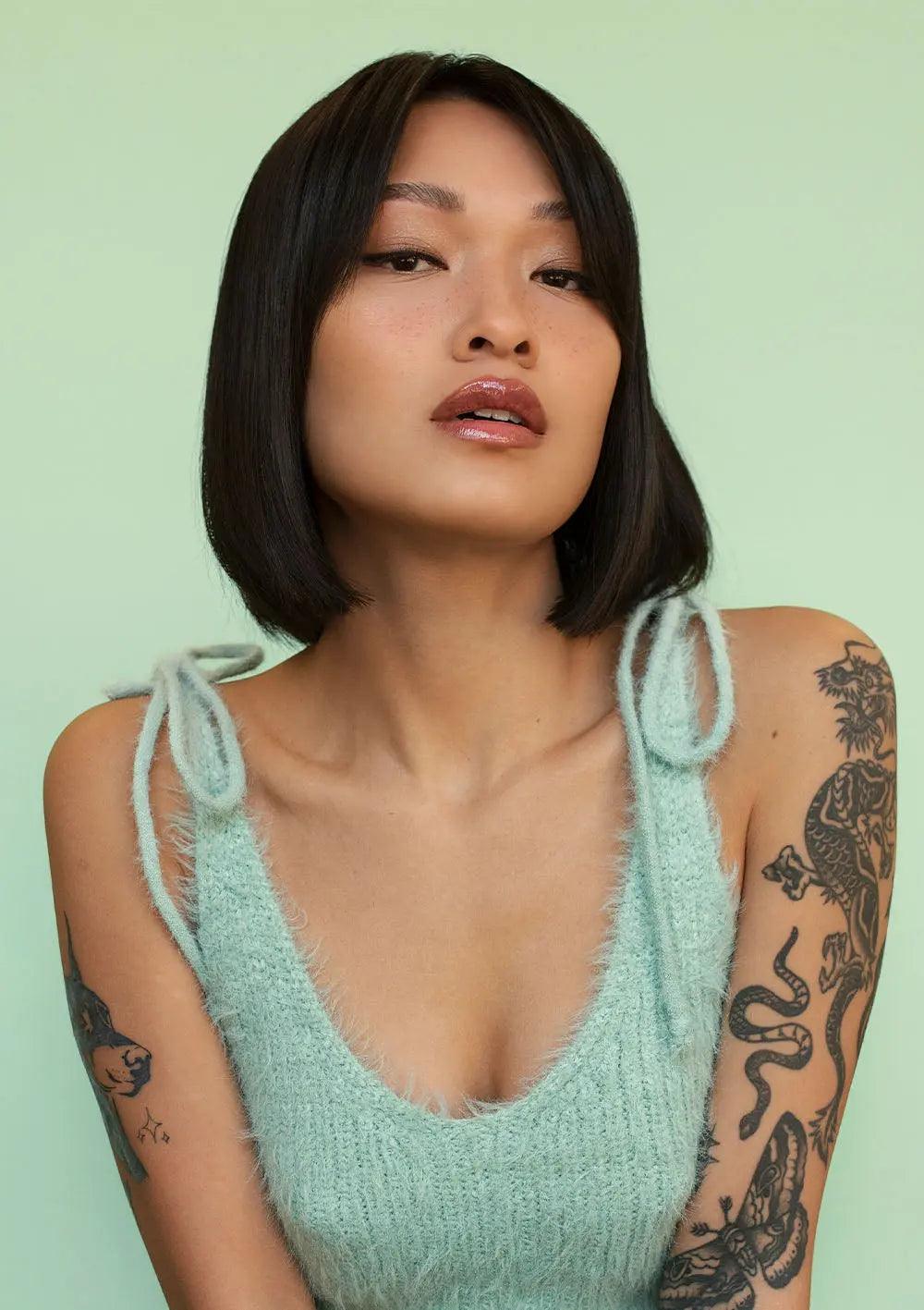 Person with tattoos wearing a light blue knit top against a mint green background, showcasing a chic bob hairstyle.