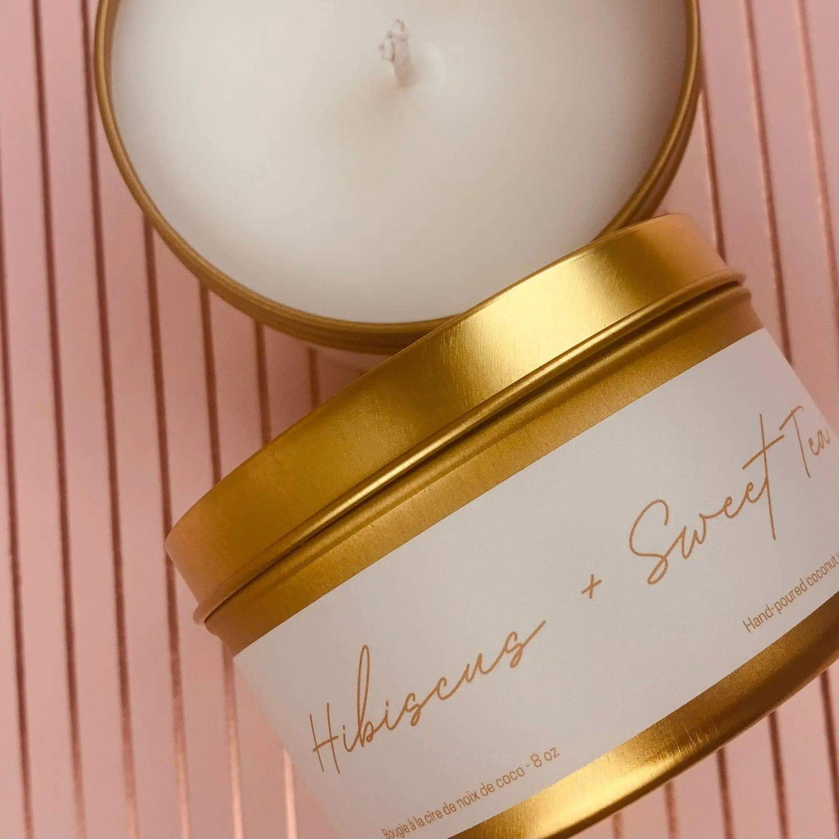 Gold tin candle labeled Hibiscus & Sweet Tea on pink striped background, made with GMO-free coconut wax and essential oils.