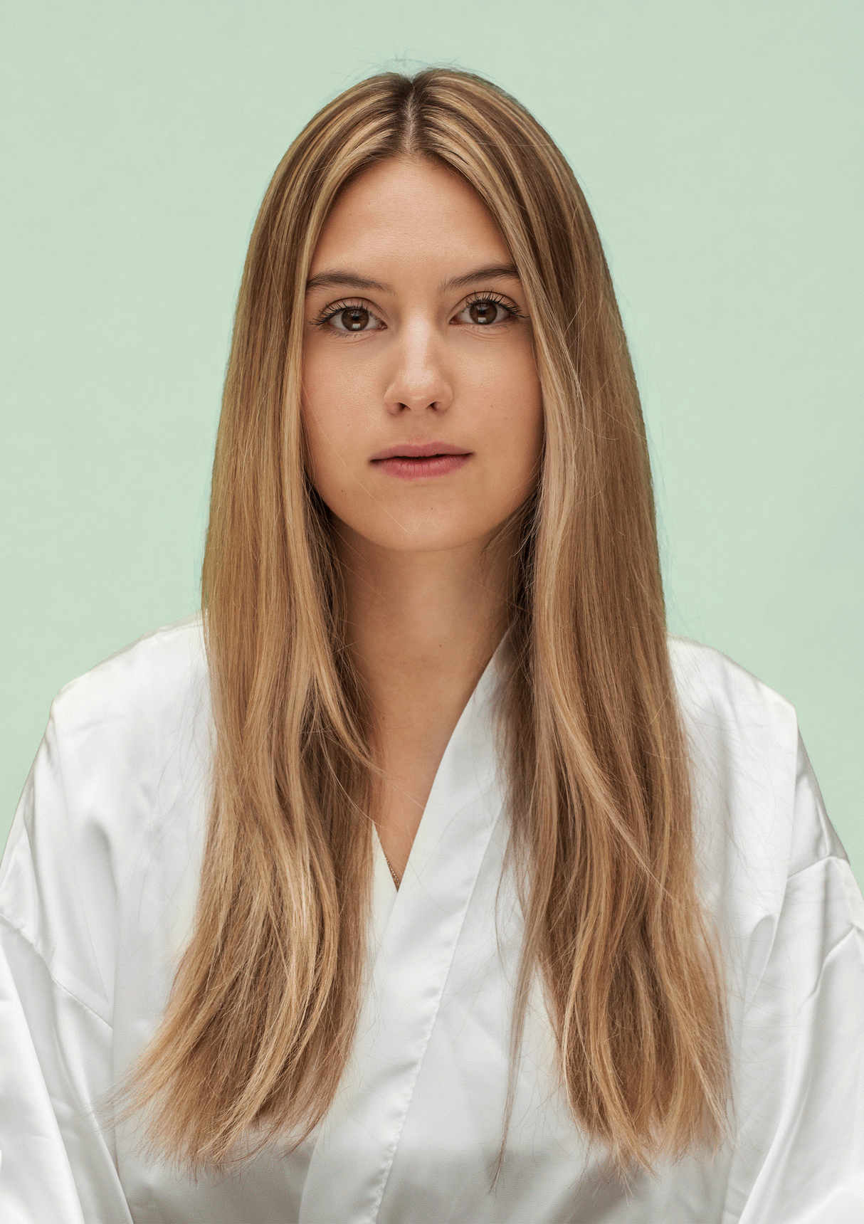 Woman with long straight hair demonstrating the effects of hydrating shampoo and conditioner, wearing a white robe against green background.
