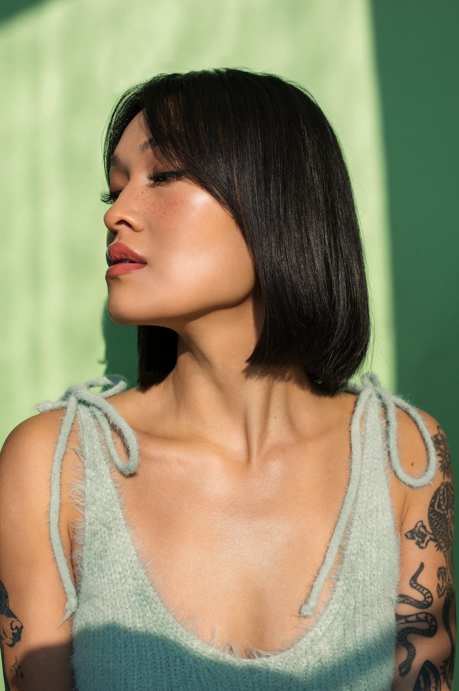 Person with shoulder-length black hair wearing a green sleeveless top, looking away from the camera with a serene expression.