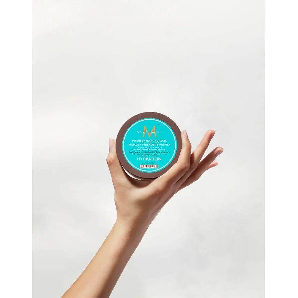 Hand holding Moroccanoil Intense Hydrating Mask jar against a plain background, highlighting product for medium to thick, dry hair hydration.