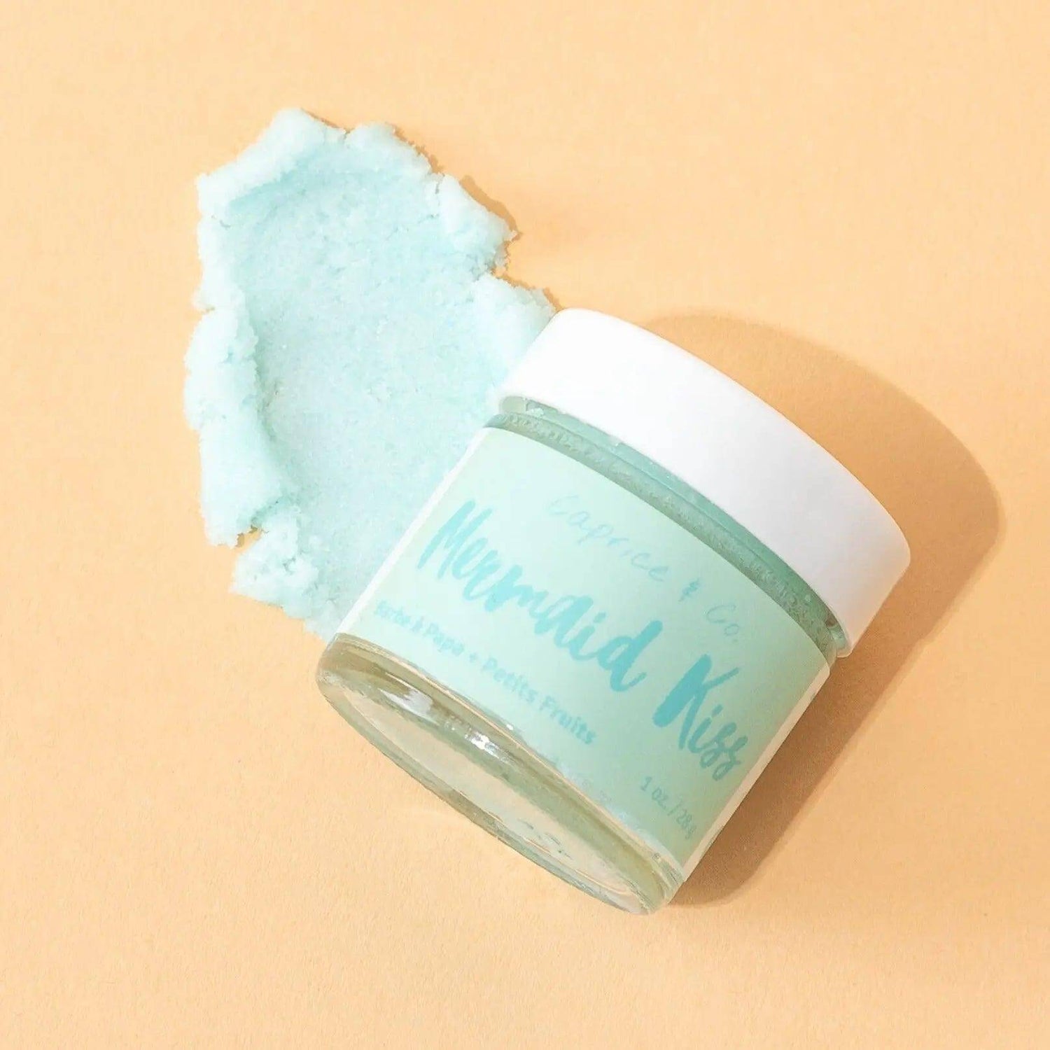 Mermaid Kiss lip scrub jar on peach background, showing light blue exfoliating texture, 1oz, cotton candy and berries scent.
