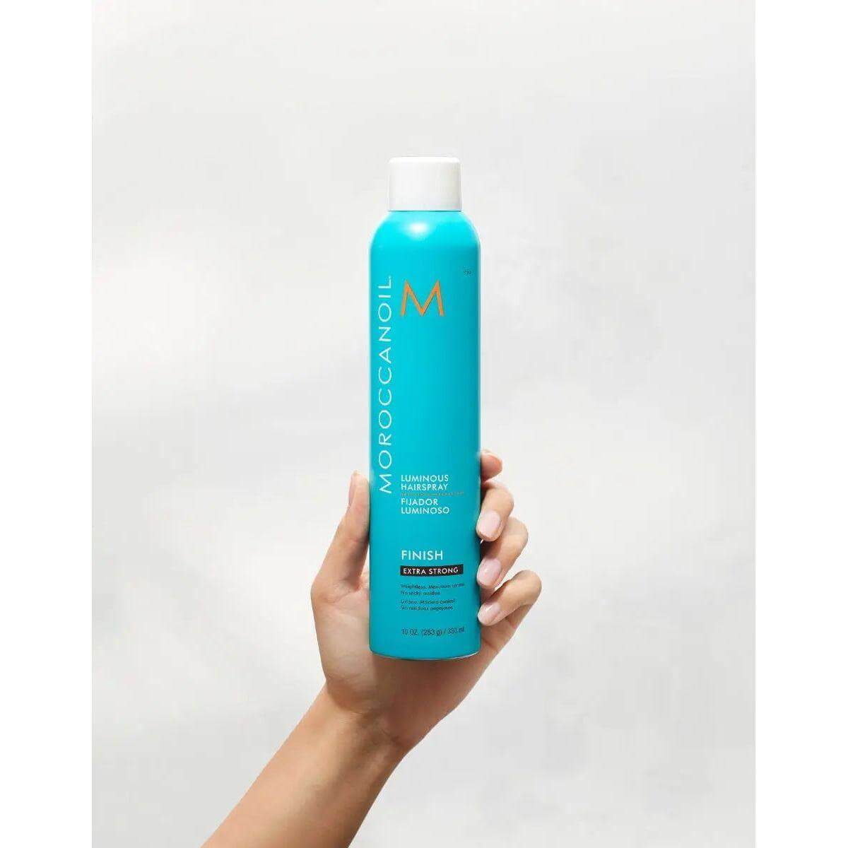 Hand holding Moroccanoil Luminous Extra Strong Finish Hairspray, 10 oz bottle, for long-lasting hold and shine.