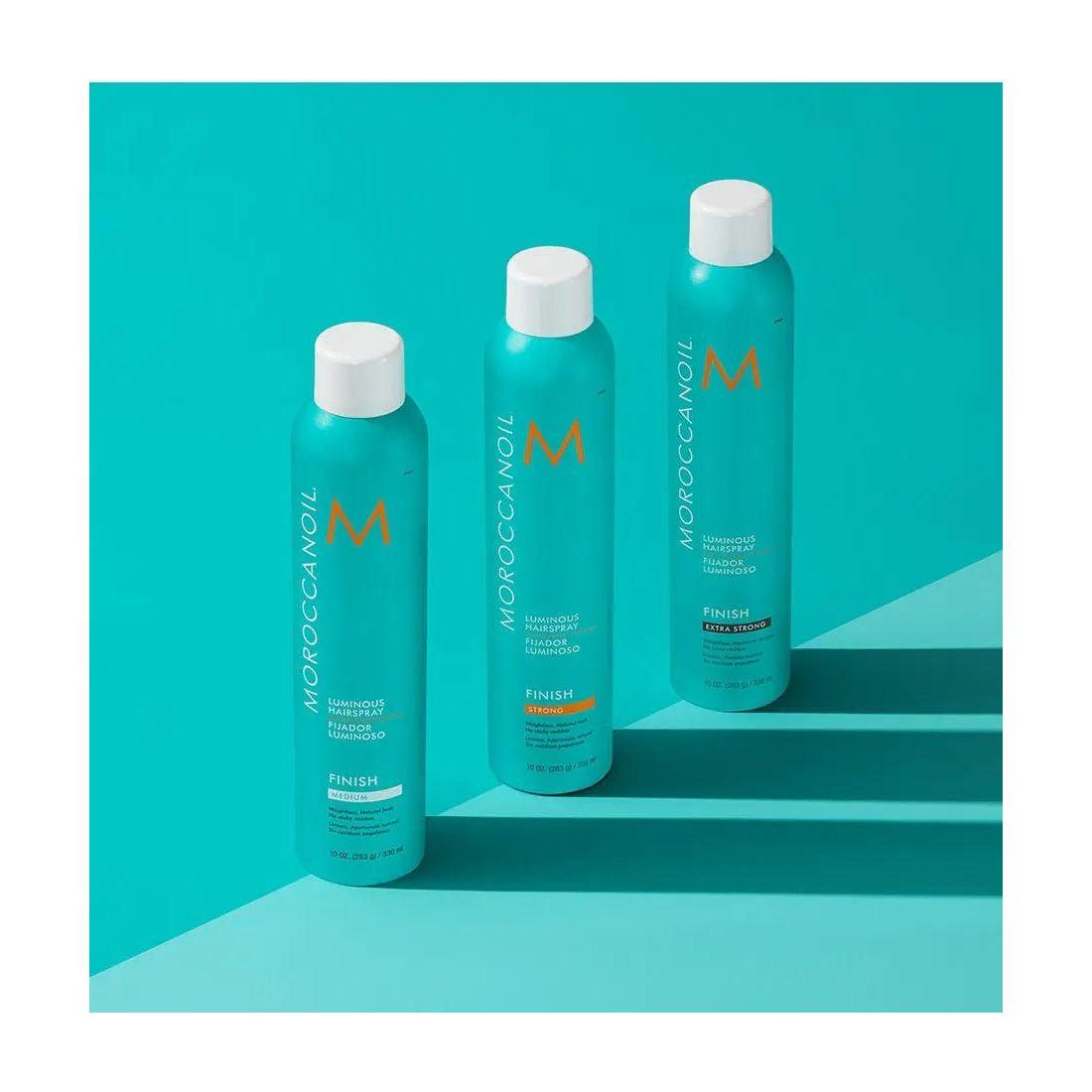 Three bottles of Moroccanoil Luminous Extra Strong Finish Hairspray on a turquoise background, showcasing refined elegance and shine.