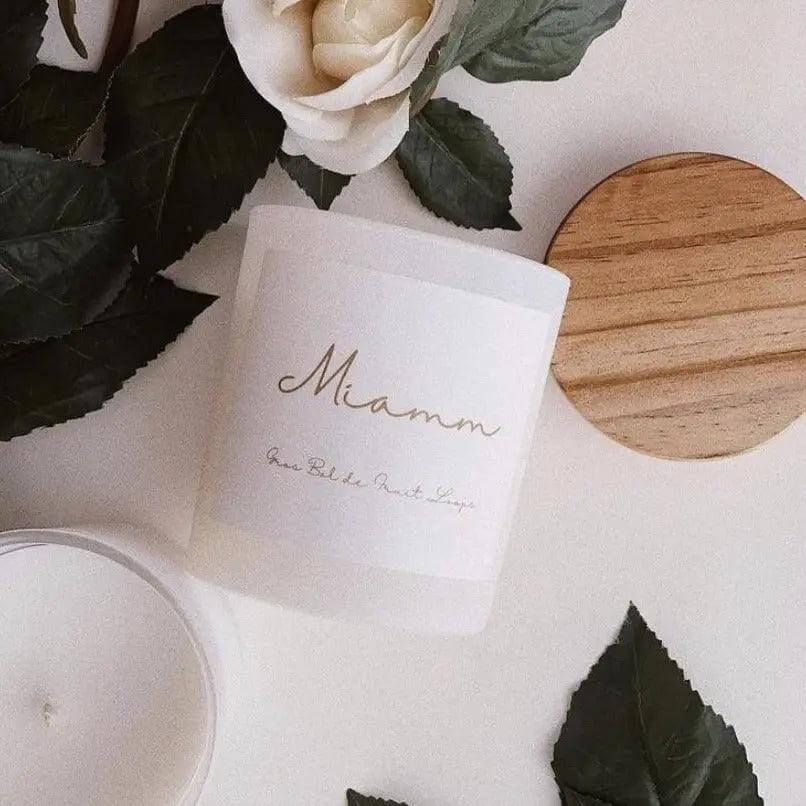 Miamm Fruit Loops candle in recyclable glass jar with wooden lid, surrounded by green leaves and a white flower.