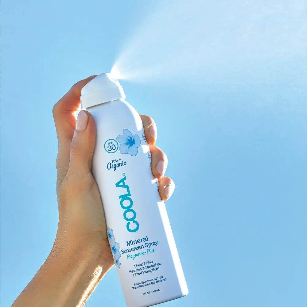 Hand holding Coola Mineral Body Organic Sunscreen Spray SPF 30, fragrance-free, against a clear blue sky.
