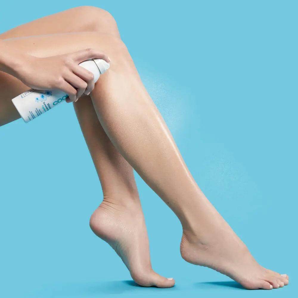 Applying fragrance-free mineral sunscreen spray SPF 30 for lightweight sun protection on legs against blue background.