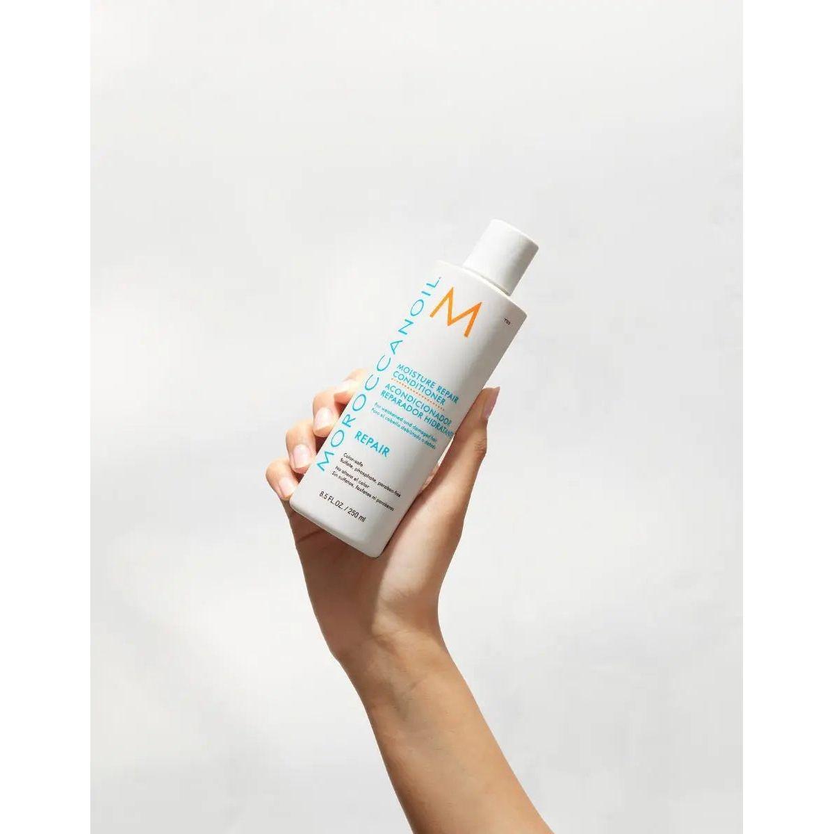 Hand holding Moroccanoil Moisture Repair Conditioner bottle against a light background.