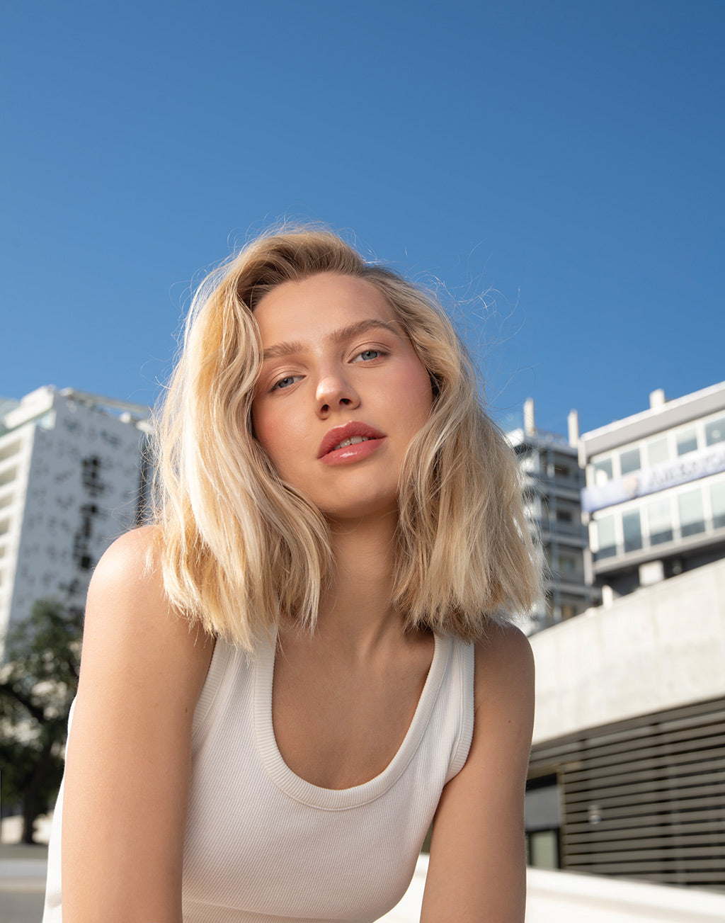 Woman with blonde hair and white tank top posing outdoors with buildings in the background.
