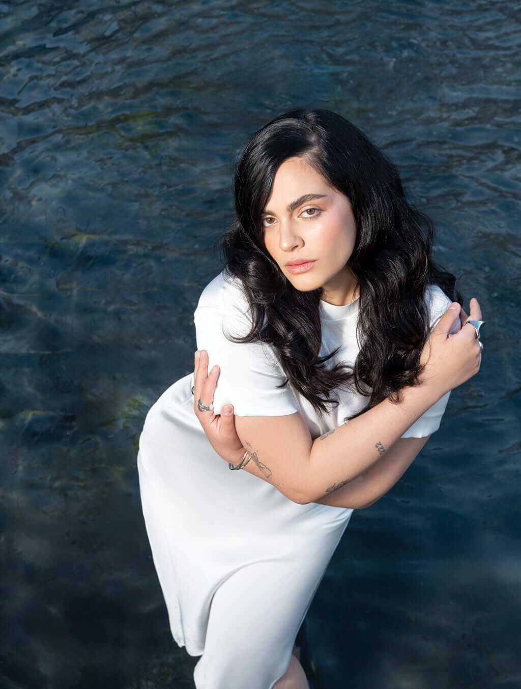 Woman standing in water wearing a white dress with long black hair, embracing herself.