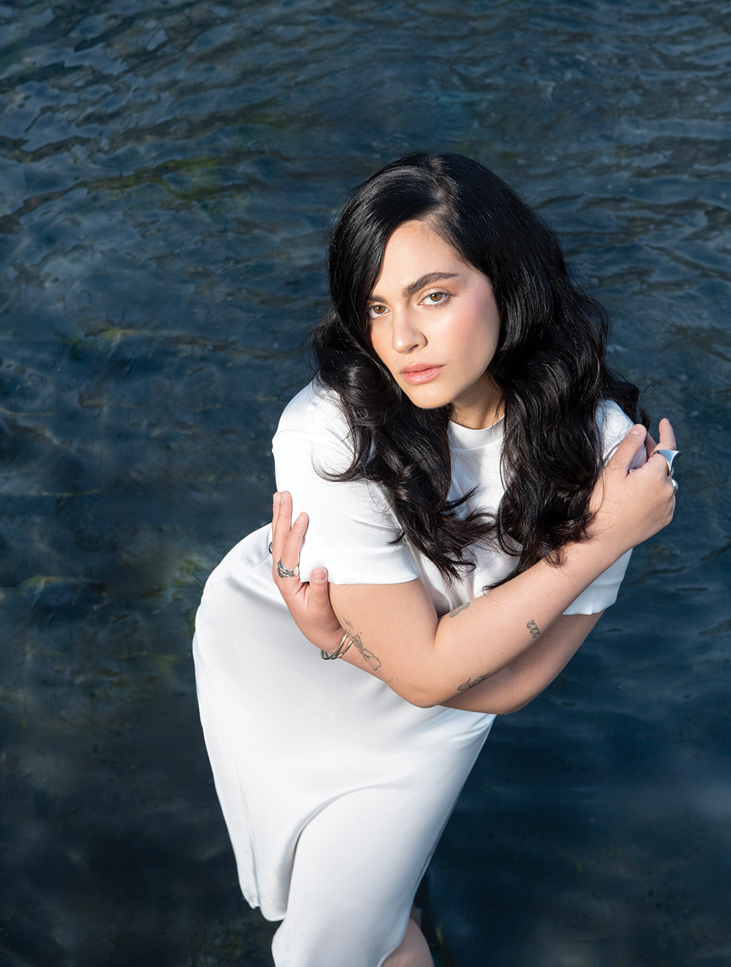 Woman in white dress with long black hair stands in water, arms crossed, looking thoughtful.