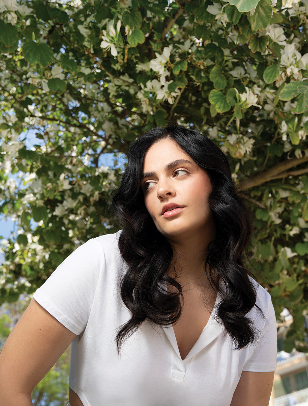Woman with long black hair and white shirt stands under a flowering tree, looking to the side.