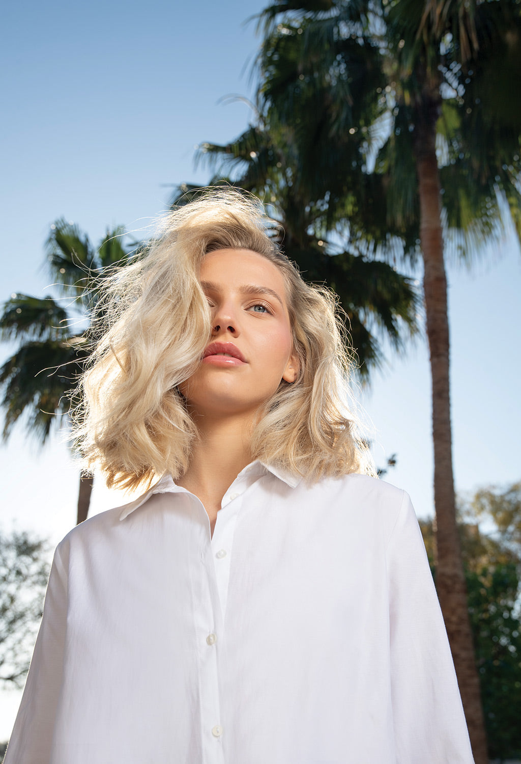 Woman with voluminous blonde hair stands outdoors, showcasing hair fullness and bounce, wearing a white shirt against a palm tree backdrop.
