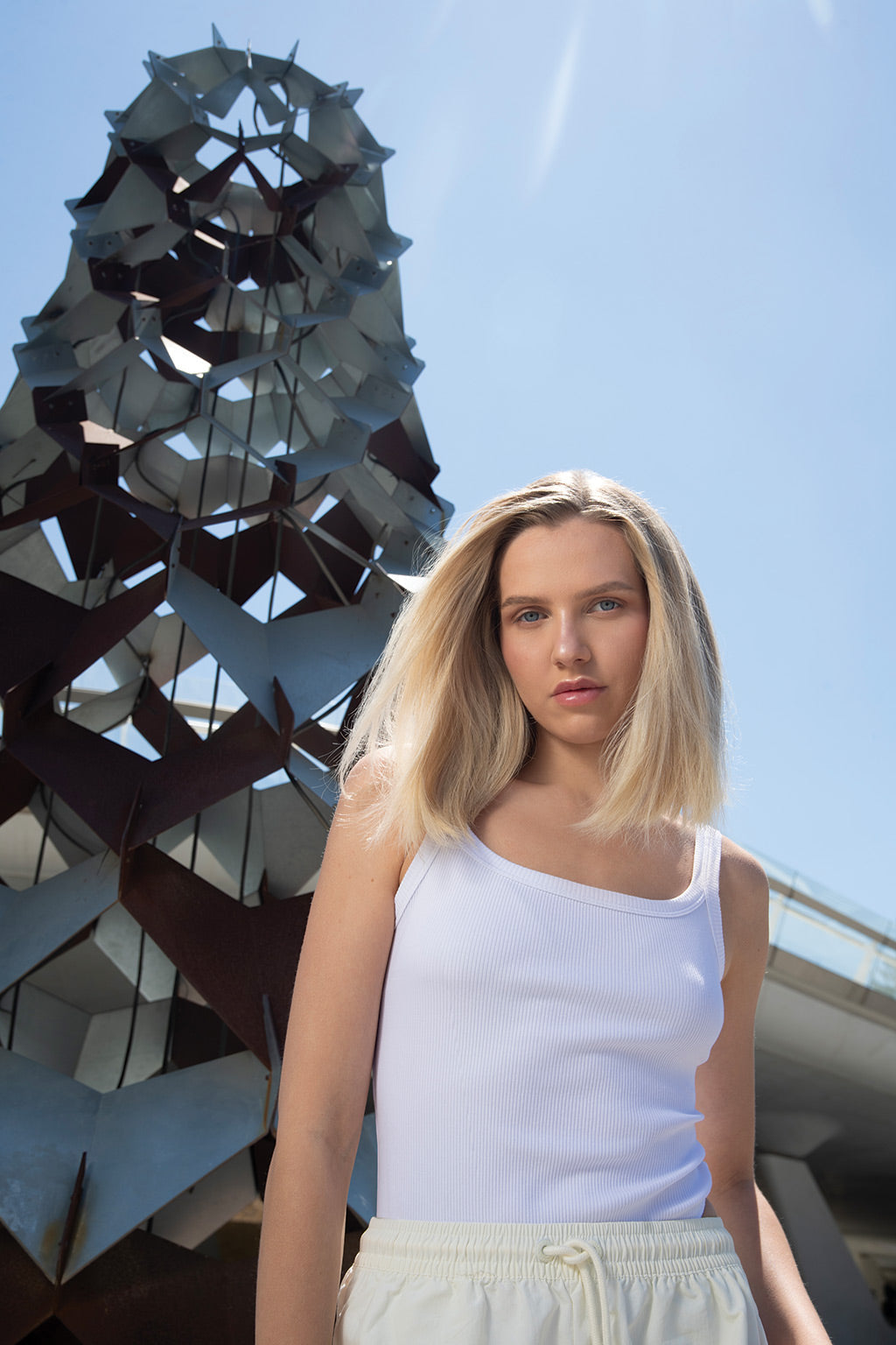 Person in white tank top standing near modern sculpture under blue sky