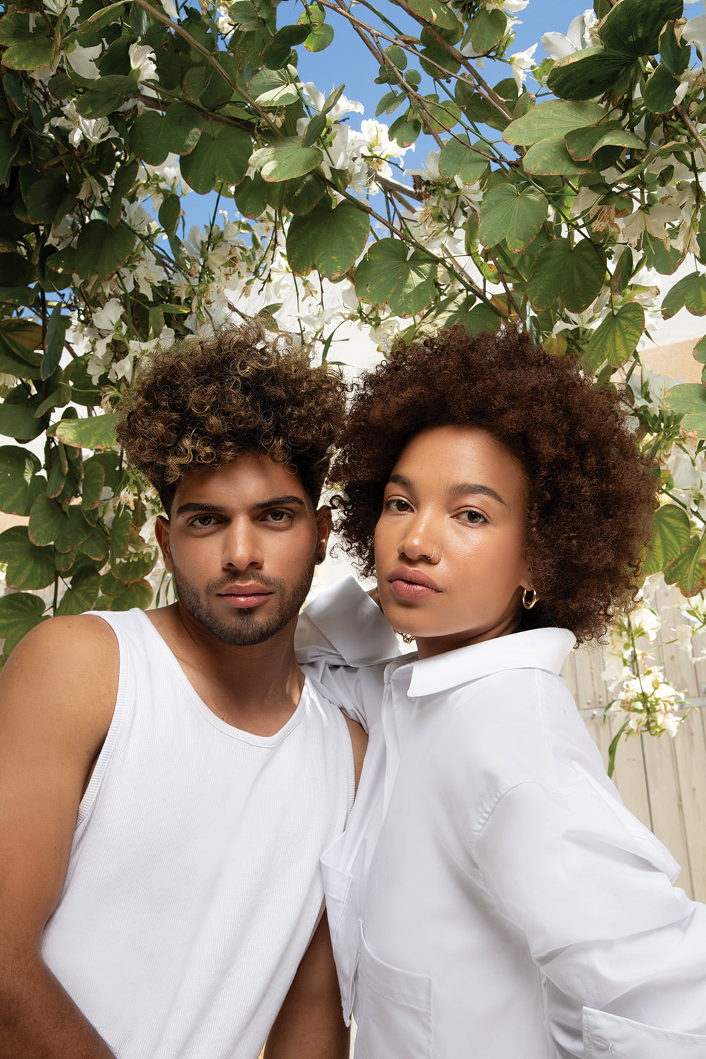 Couple with curly hair posing outdoors with blooming flowers in the background.