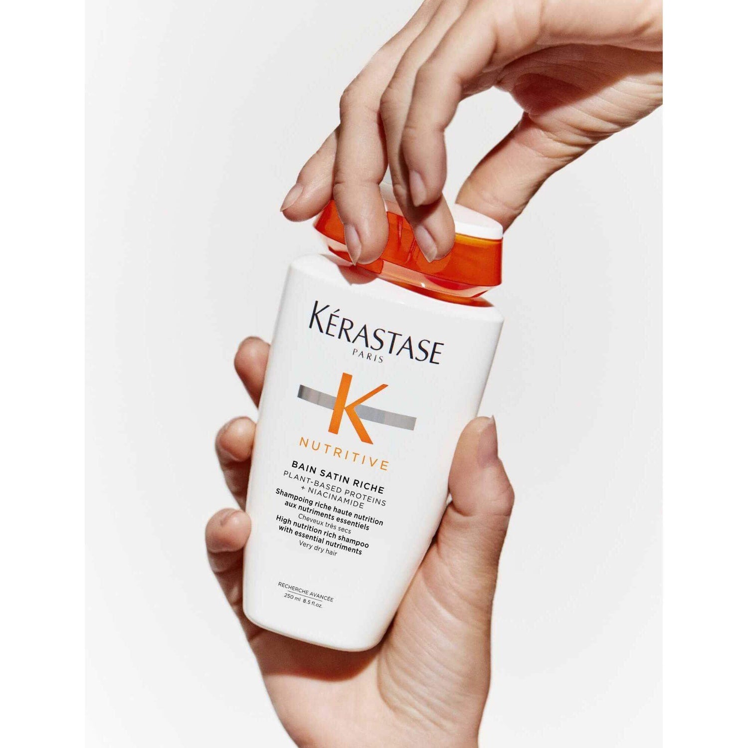 Hands holding Kérastase Nutritive Bain Satin Riche Shampoo bottle for very dry hair nourishment.
